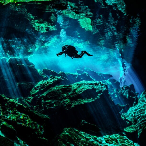 Eine Person in Taucherausrüstung taucht in einer von Wasser gefüllten Höhle. Lichtsäulen ergeben sich durch den starken Lichteinfall von oben im Wasser.