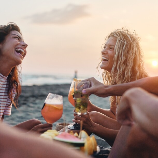 Eine Gruppe von vier fröhlichen Personen sitzt am Meer bei Sonnenuntergang mit Drinks und Snacks.