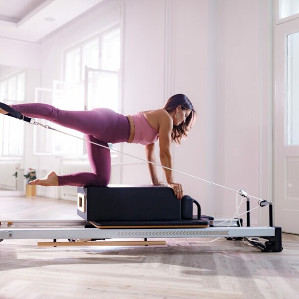 Eine Frau in pinker Sportkleidung kniet auf einer Box auf einem Pilates Reformer in einem hellen Raum.