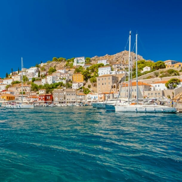 Mediterrane Ortschaft in Hügellage mit Hafenpromenade, im Vordergrund türkisblaues Meer mit Booten.