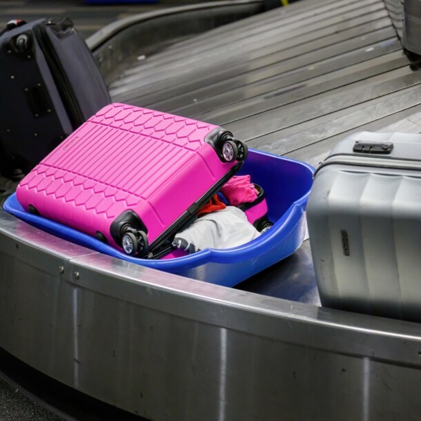 Ein pinker Hartschalenkoffer liegt geöffnet in einer Plastikwanne auf einem Gepäckband neben anderen Koffern.
