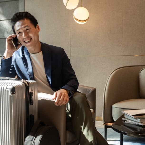 Ein fröhlicher asiatischer Geschäftsmann sitzt im Sessel einer modernen Flughafen-Lounge und telefoniert an seinem Smartphone, neben ihm steht ein großer Reisekoffer.