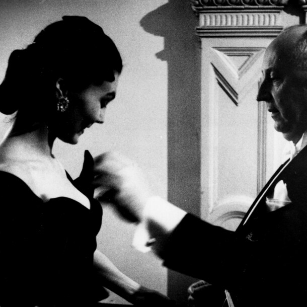 Profilansicht von Christian Dior in einem schwarzen Anzug, der etwas an einem schwarzen Abendkleid eines weiblichen Models stylt, Schwarzweißfoto.