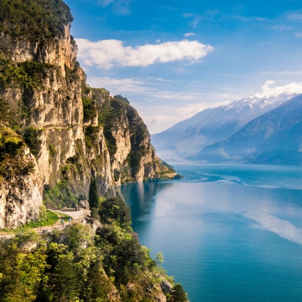 Bewachsene Felswand nahe Riva del Garda direkt am Gardasee mit Gipfeln auf der anderen Seeseite im Hintergrund.