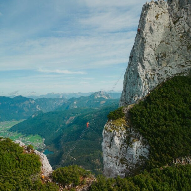 Eine Person auf einer Leiter, die hoch über einem Abgrund zu einem hohen Felsen führt, im Hintergrund Berge und Himmel.