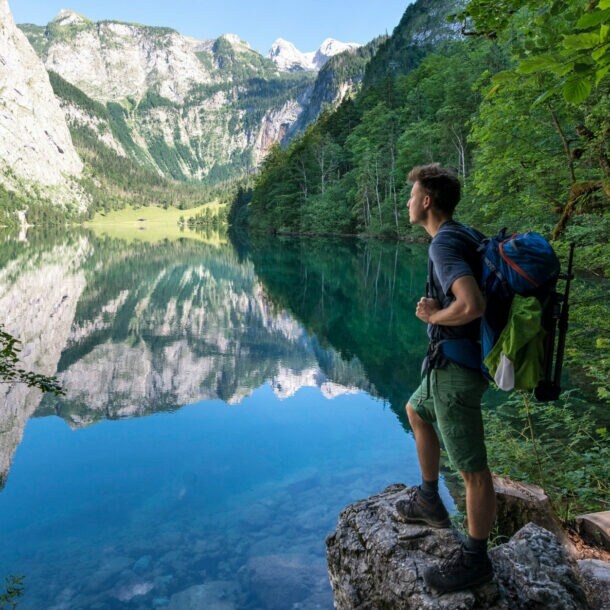 Eine Person mit Wanderrucksack steht auf einem Stein vor dem Königssee mit Blick auf die Berge.