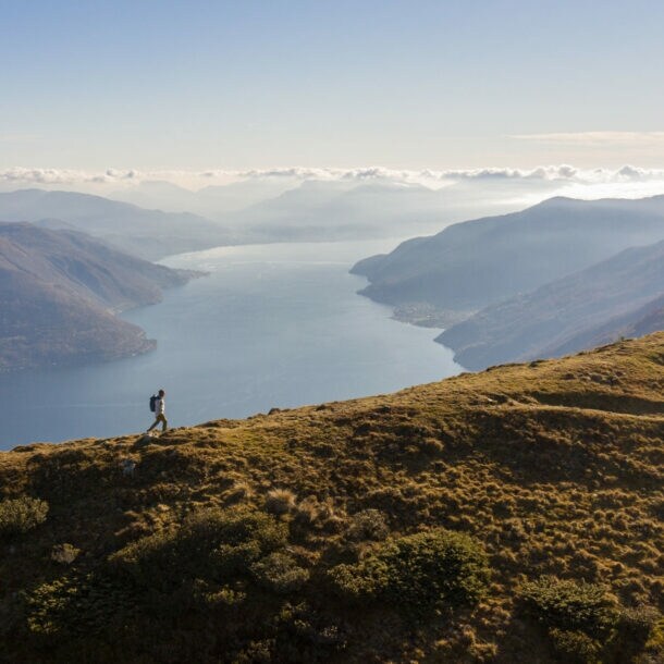 Panoramaaufnahme von einem Mann mit Rucksack auf einem Berg und dem Lago Maggiore im Hintergrund.