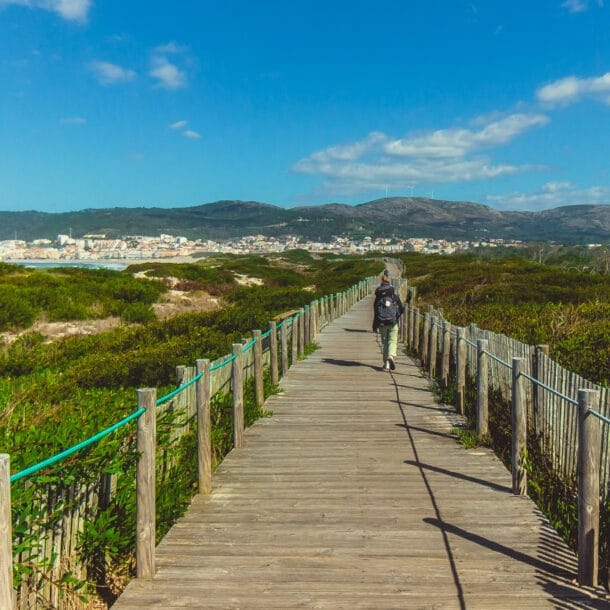 Eine unkenntliche Person beim Wandern in Portugal auf einem Holzsteg, der in Küstennähe über grüne Vegetation führt.