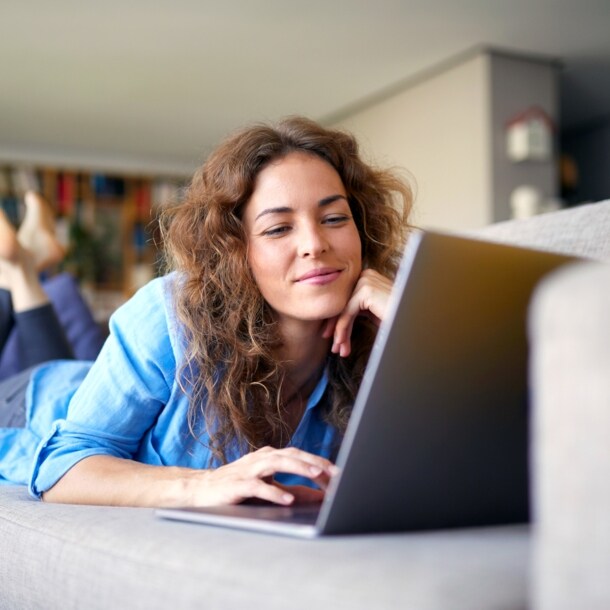 Eine Frau liegt bäuchlings auf einem Sofa in einem Wohnzimmer und schaut lächelnd auf einen Laptop vor ihr.