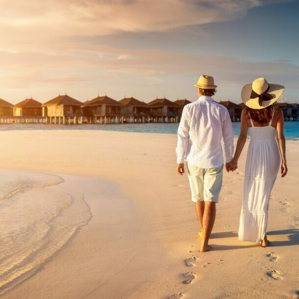 Rückansicht eines händchenhaltenden Paares in heller Sommerkleidung und mit Strohhüten an einem Strand vor einem Holzstegmit Wasserbungalows.