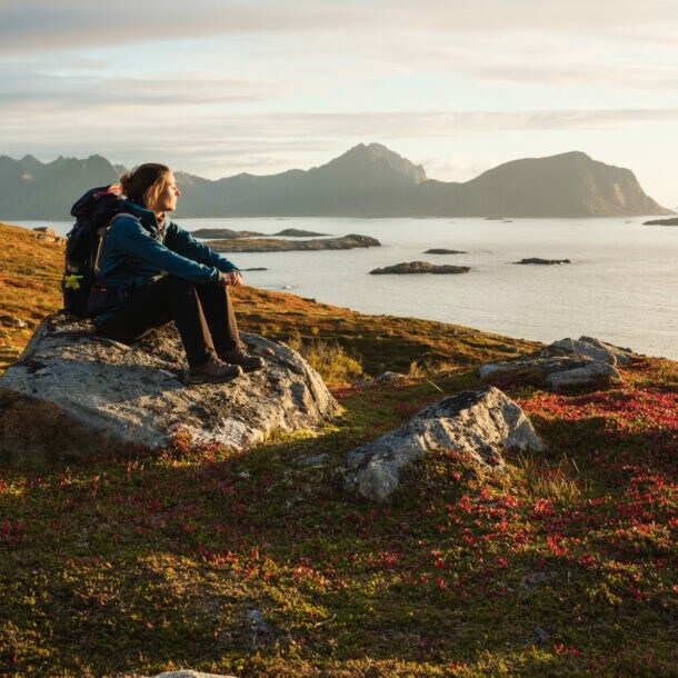 Eine Frau mit Wanderausrüstung, die bei Sonnenuntergang auf einem großen Stein sitzt und aufs Wasser blickt, im Hintergrund Berge.
