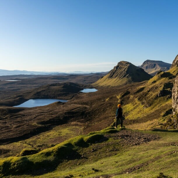 Eine Person steht in einer Landschaft aus Wiesen, Hügeln und Seen in Schottland. 