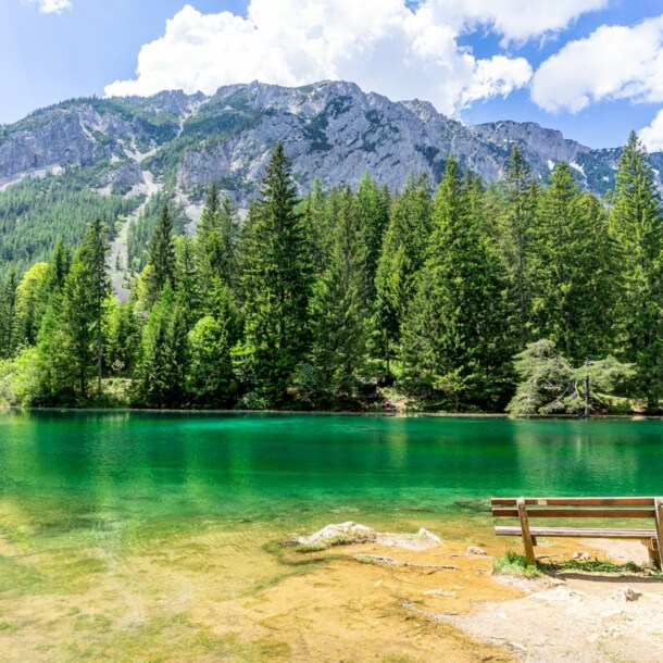 Eine Sitzbank vor einem grün schimmernden See, umgeben von Nadelwald in einer Bergregion.