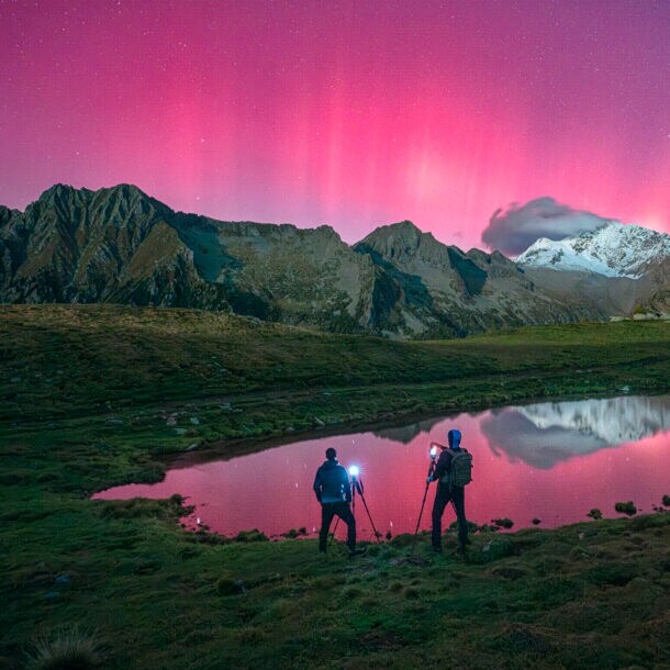 Zwei Menschen stehen vor einer Bergkulisse und fotografieren Polarlichter, die den Himmel pink färben.