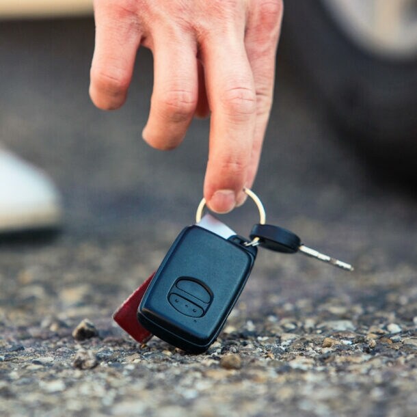 Nahaufnahme einer Hand, die nach einem Autoschlüssel am Asphaltboden greift, dahinter ein Autoreifen und Sneaker in Unschärfe.