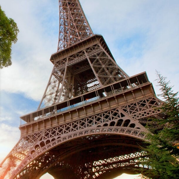 Mittelteil des Eiffelturmes mit Personen auf einer Aussichtsplattform, im Vordergrund grüne Bäume.