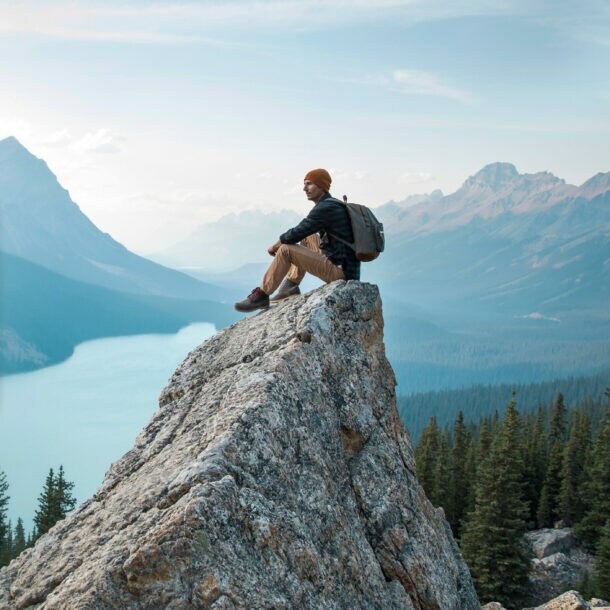 Ein Mann mit Rucksack sitzt auf einem spitzen Felsen in einer Fjordlandschaft mit Nadelwald.