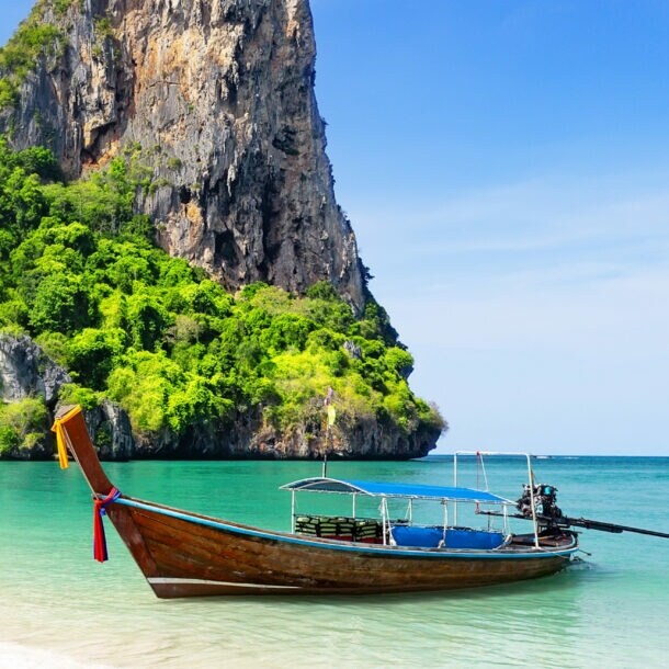Ein Longtail-Boot im smaragdgrünen Wasser an einem weißen Sandstrand vor einem begrünten Felsen.