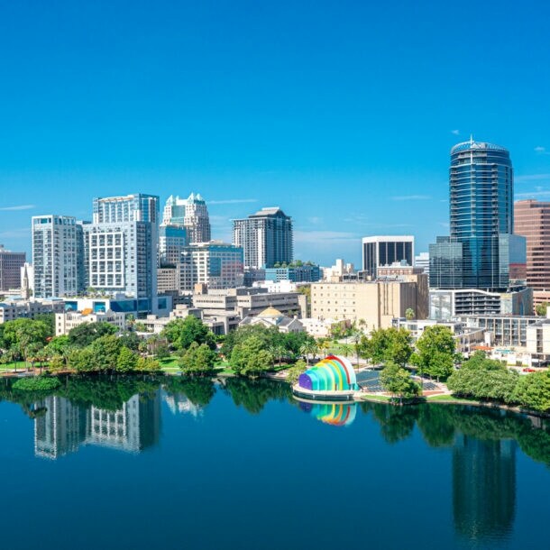 Skyline von Orlando mit Hochhäusern hinter einem See mit Parkanlage unter blauem Himmel.