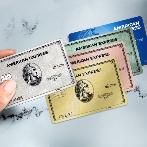 Eine Hand hält eine American Express Platinum Kreditkarte über drei weitere Kreditkarten in unterschiedlichen Farben auf einem Marmortisch.