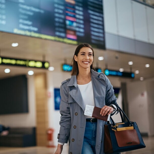 Junge lächelnde Frau in taubenblauem Trenchcoat im Abflugbereichs eines Flughafens.