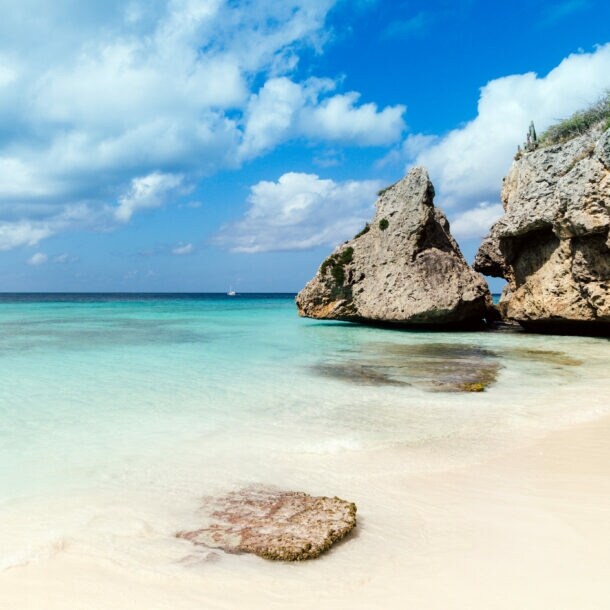 Weißer Strand am türkisblauen Meer, am Rand große Felsen im Wasser.