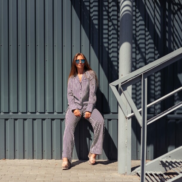 Junge Frau mit Sonnenbrille in blau-weiß gestreiftem Pyjama lehnt an eine Wand aus grauem festem Wellblech.