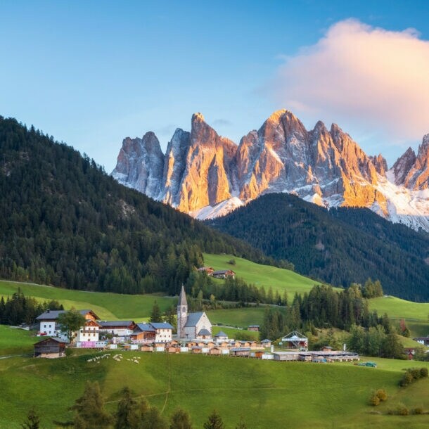 Blick auf das idyllische Dorf Sankt Maddalena, eingebettet zwischen grünen Hügeln, Wäldern und den markanten Geislerspitzen im Hintergrund.