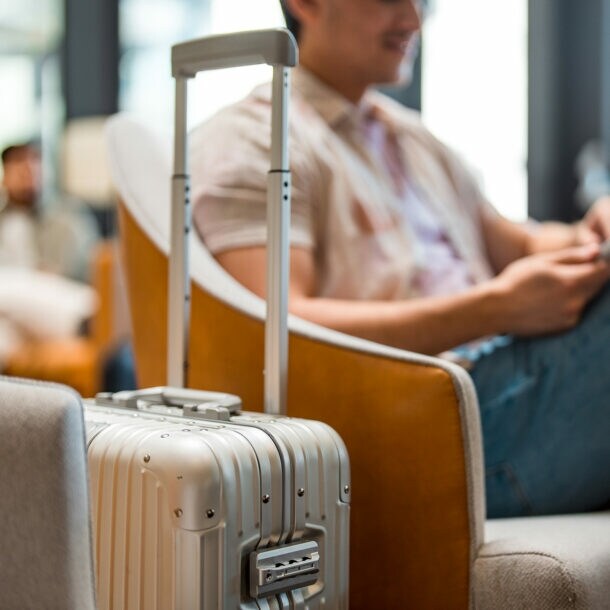 Ein Mann sitzt im Wartebereich eines Flughafens. Im Vordergrund steht sein Rollkoffer.