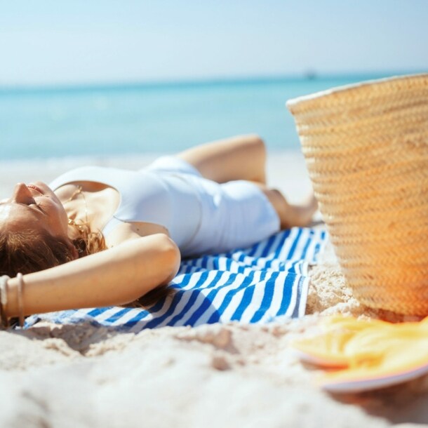 Eine Frau liegt rücklings entspannt auf einem gestreiften Badehandtuch an einem Sandstrand am Meer, neben ihr ein Strohkorb und Badeschuhe.