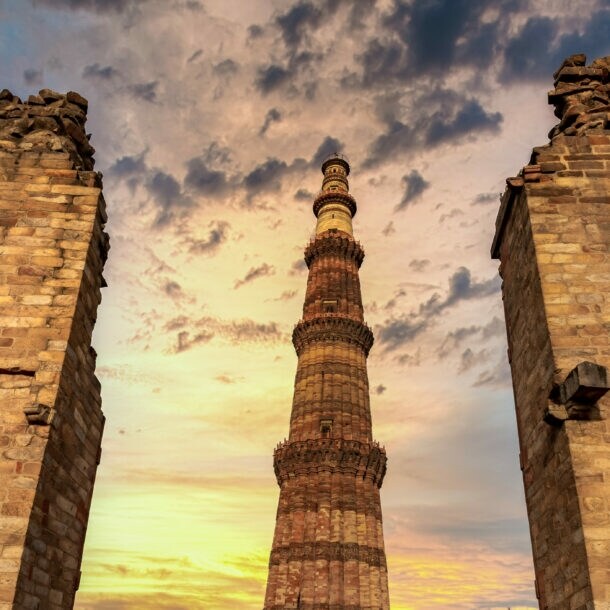 Das Qutub Minar, ein hoher roter Sandsteinturm, umgeben von Ruinenmauern bei Sonnenuntergang mit bewölktem Himmel