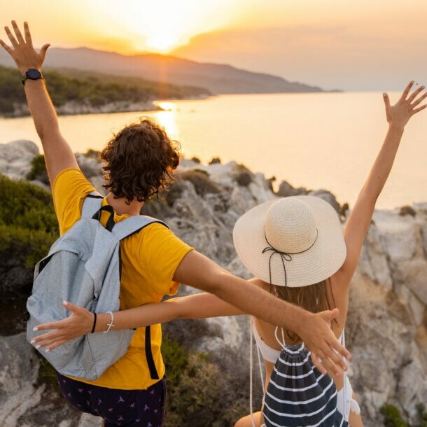 Ein Paar steht mit offenen Armen auf einem Felsen und schaut auf das Meer und den Sonnenuntergang.