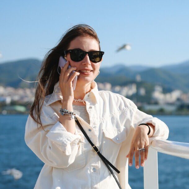 Eine Frau mit Sonnenbrille und wehenden Haaren telefoniert mit einem Smartphone an der Reling eines Schiffes auf dem Meer, im Hintergrund fliegen Möwen vor Stadtpanorama an einer Küste.