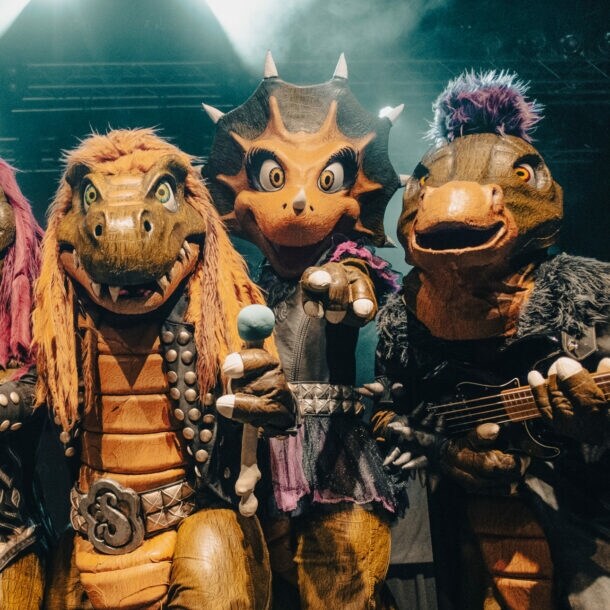 Vier Personen in Dinosaurierkostümen, die in die Kamera gucken und teilweise Instrumente und ein Mikrofon halten