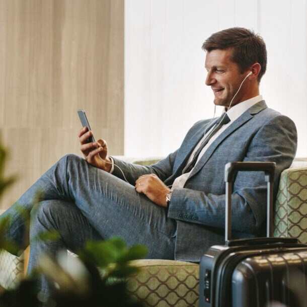 Ein Geschäftsmann sitzt in einem Sessel in einer Flughafen-Lounge und schaut lächelnd auf sein Smartphone.