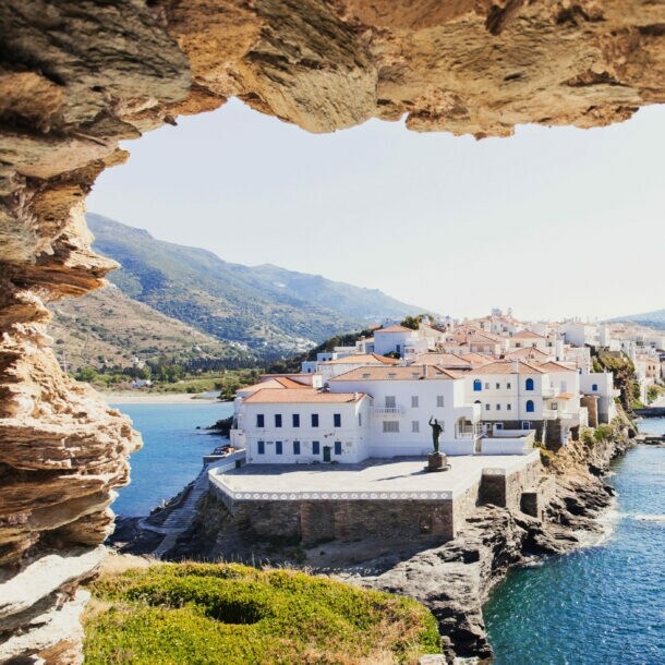 Blick auf weiße Häuser eines Ortes auf einer felsigen Landzunge im Meer durch ein Loch in einer Felsenmauer.
