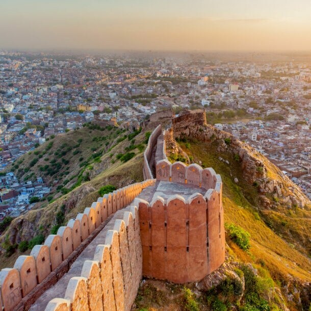 Luftaufnahme der historischen Stadtmauer von Jaipur, die sich über grüne Hügel erstreckt, mit der Stadt im Hintergrund bei Sonnenuntergang