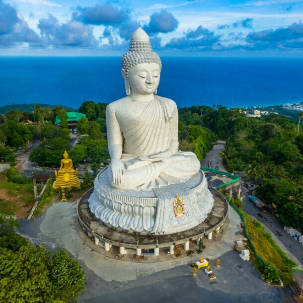 Eine große weiße Buddha-Statue auf einem bewaldeten Hügel an einer Meeresküste.