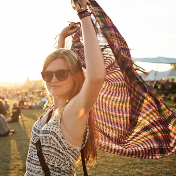Eine Frau mit Sonnenbrille steht auf einer Wiese, auf der Leute sitzen, und hält ein Tuch über ihren Kopf.