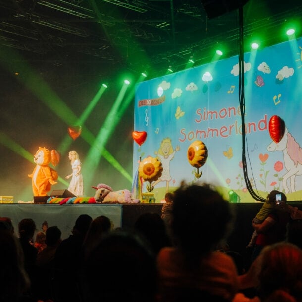 Simone Sommerland steht auf einer Bühne voller Luftballons, neben ihr eine Person im Eichhörnchen-Kostüm, vor ihr Publikum.
