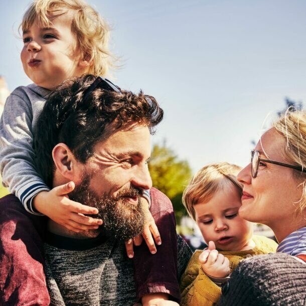 Vater trägt lachendes Kleinkind auf Schultern, Mutter mit Brille schaut liebevoll, während sie ein weiteres Kleinkind im Arm hat.