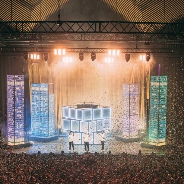 Große Menschenmenge bei einem Konzert oder Event in einer Halle mit beleuchtetem Bühnenbereich und Nebeleffekten.