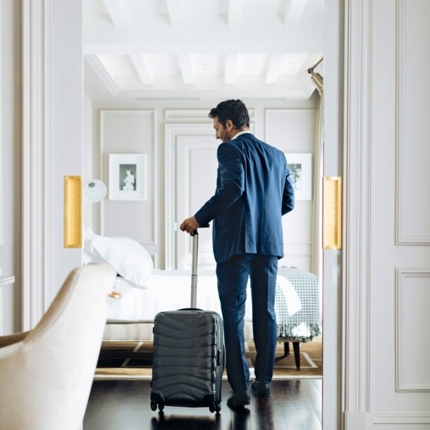 Mann im Anzug und einem Rollkoffer in der Hand betritt eine helle luxuriöse Hotel-Suite.