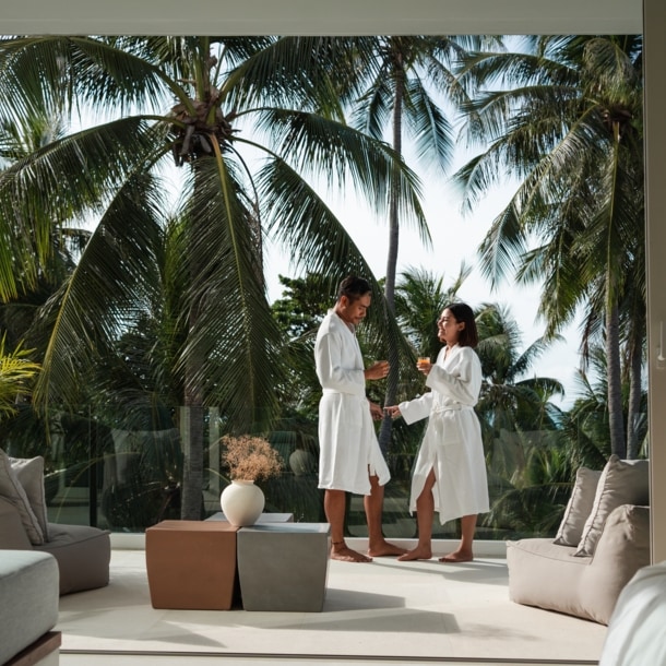 Ein glückliches Paar in weißen Bademänteln steht auf einem, von grünen Palmen umgebenen Hotelbalkon und hält ein Glas Saft in den Händen.
