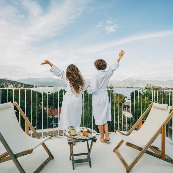 Zwei Menschen in weißen Bademänteln von hinten stehen auf einem Balkon, blicken ins Grüne und strecken freudevoll die Arme in die Luft.