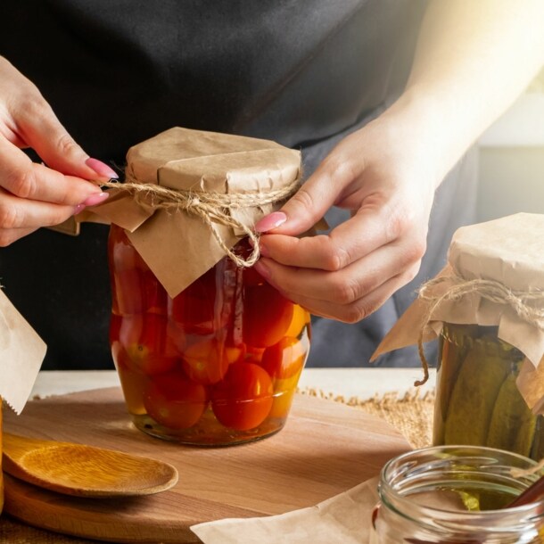 Person bindet eine Schnur um den Deckel eines Einmachglases mit eingelegtem Gemüse auf einem Holztisch.