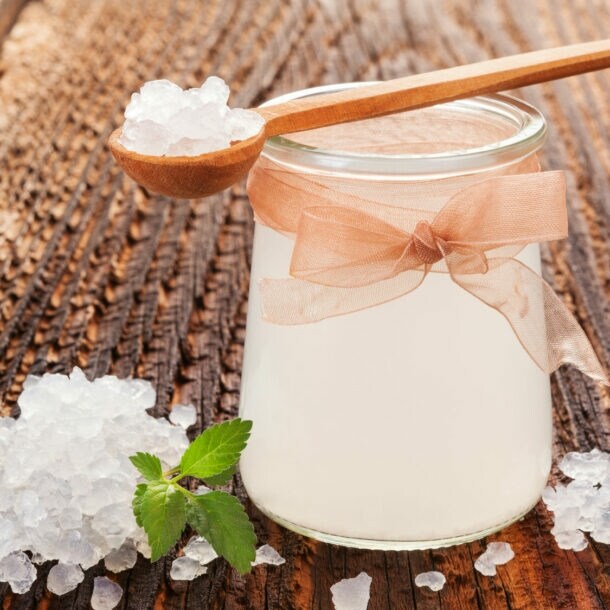 Glas mit milchiger Flüssigkeit und Schleife, umgeben von kristallartigen weißen Körnern auf Holzoberfläche