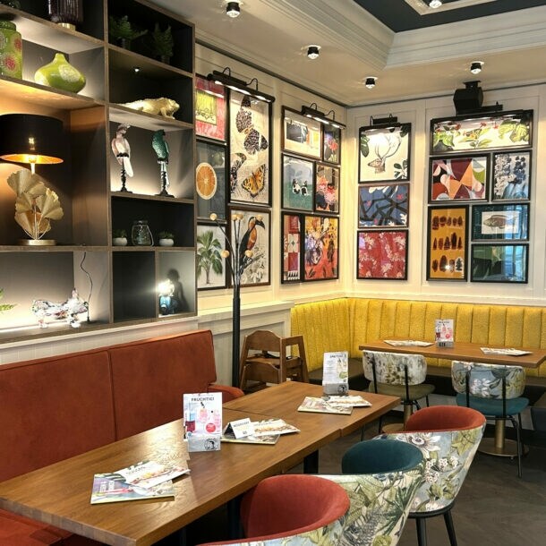 Gemütlicher Gastraum mit gelber Eckbank, bunten Stühlen und zahlreichen farbenfrohen Bildern an den Wänden.