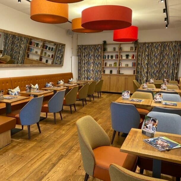 Gastraum mit Holzboden, bunten Stühlen, brauner Polsterbank, großen farbigen Deckenlampen und Bücherregal im Hintergrund.