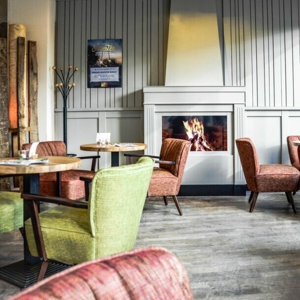 Gemütlicher Gastraum des ALEX in Wiesbaden mit loderndem Gaskamin, runden kleinen Tischen und bequemen Sesseln in Grün und Rot.